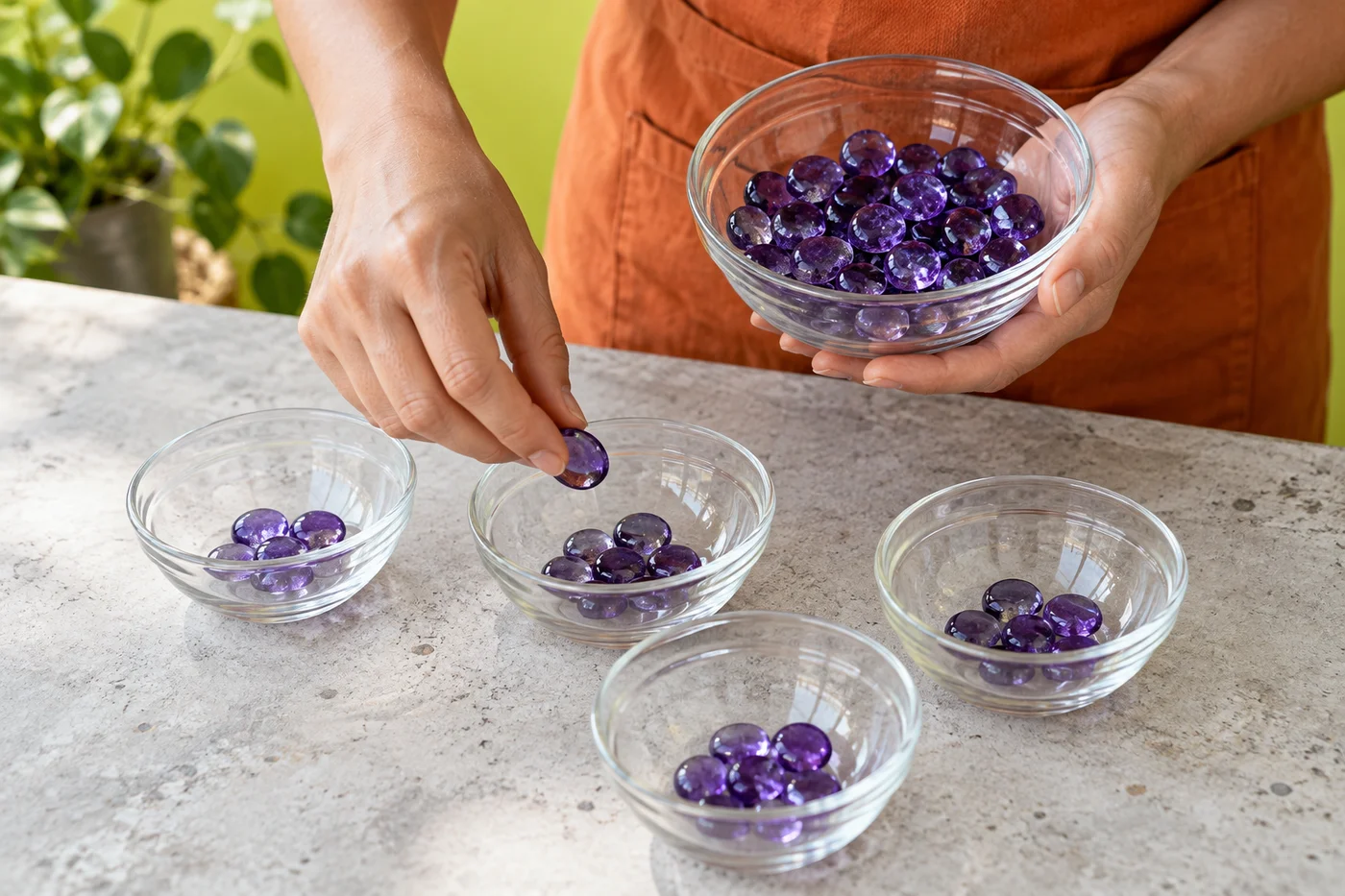 Erwachsene Hände verteilen violette Glassteine auf klare Schalen auf einer hellen Werkbank