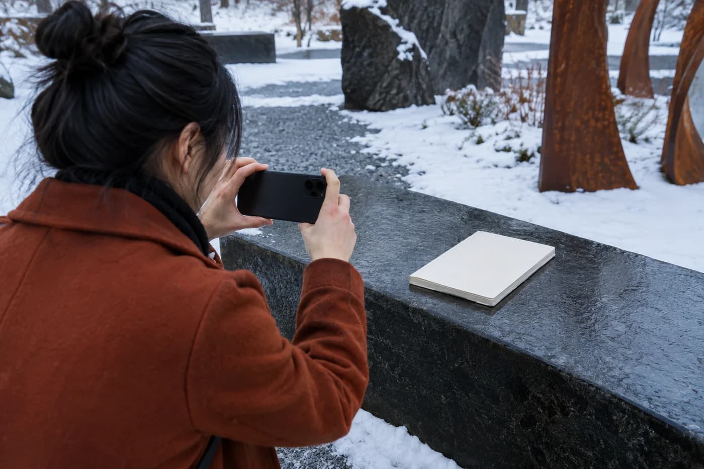 Mujer adulta con abrigo color óxido sosteniendo un teléfono sobre un cuaderno entero en blanco en un banco nevado