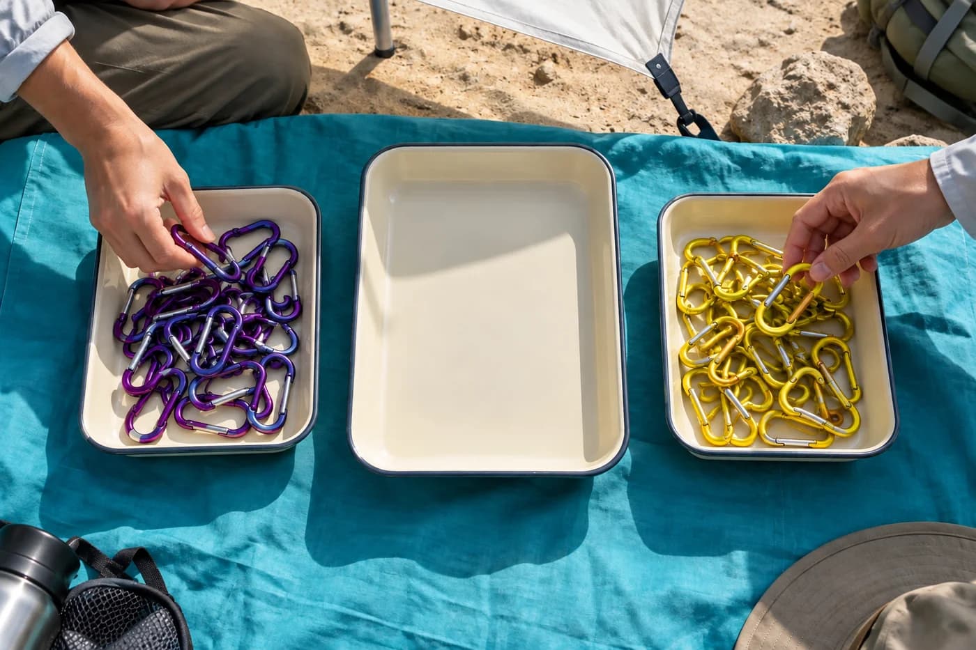 Adult hands choosing violet and yellow metal clips from two cream trays beside an empty larger tray on cyan fabric