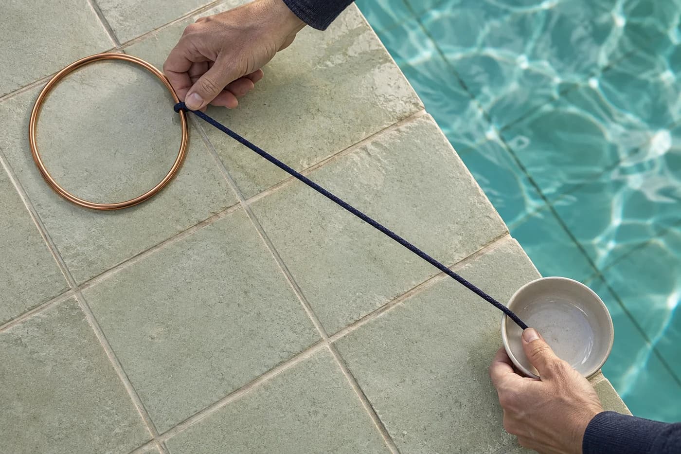 Erwachsene Hände spannen eine marineblaue Schnur zwischen Kupferreifen und Keramikschale auf einem trockenen gefliesten Poolrand