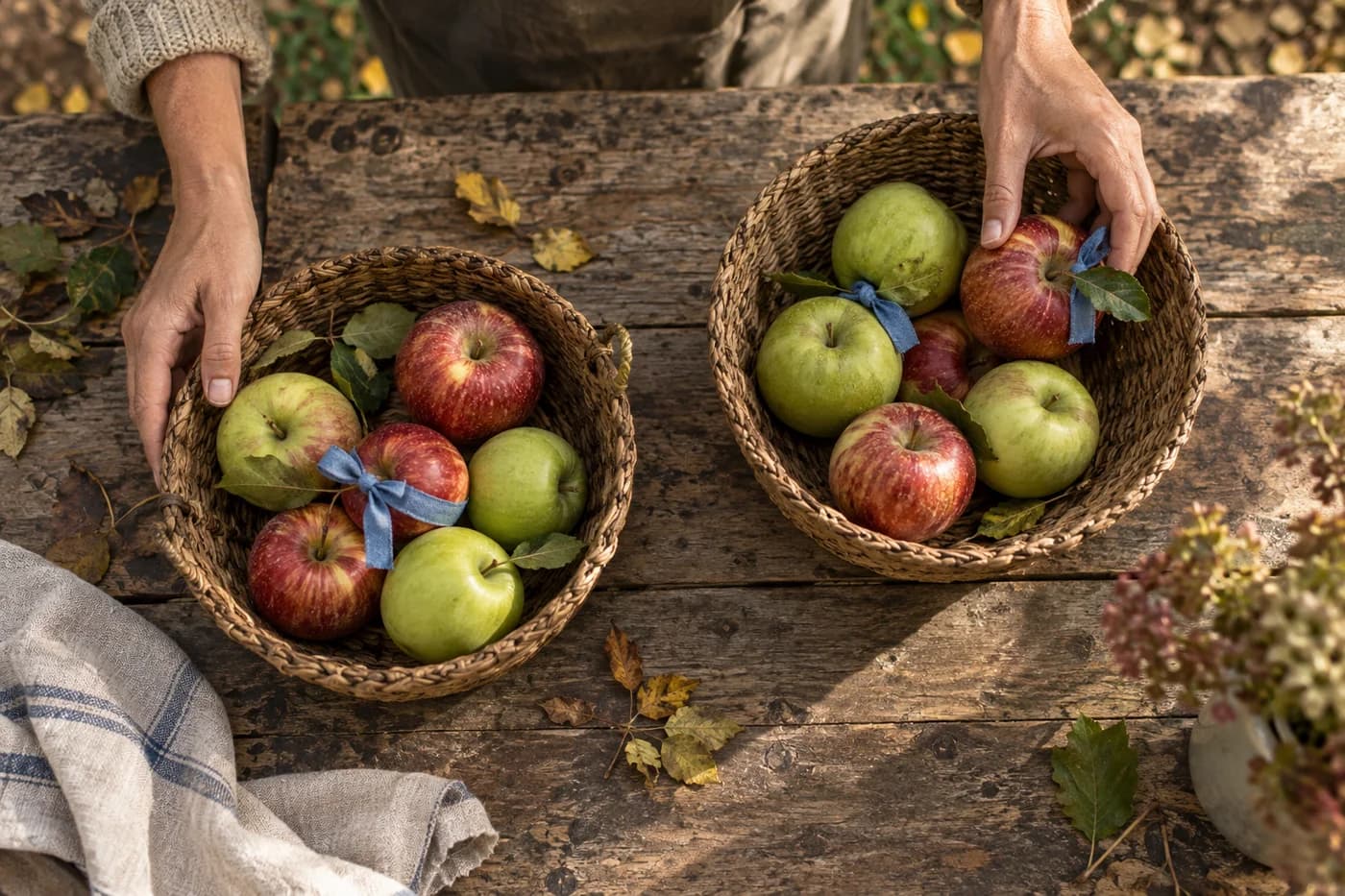 Two woven baskets of mixed red and green apples with blue linen ribbons on several apples, viewed from above