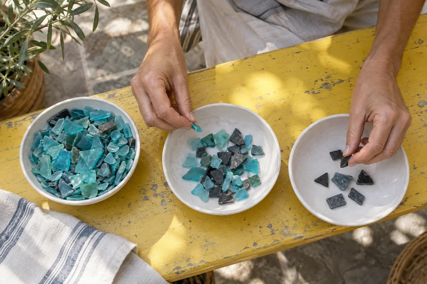 Adult hands sorting mosaic fragments into differently filled shallow white bowls on a yellow bench
