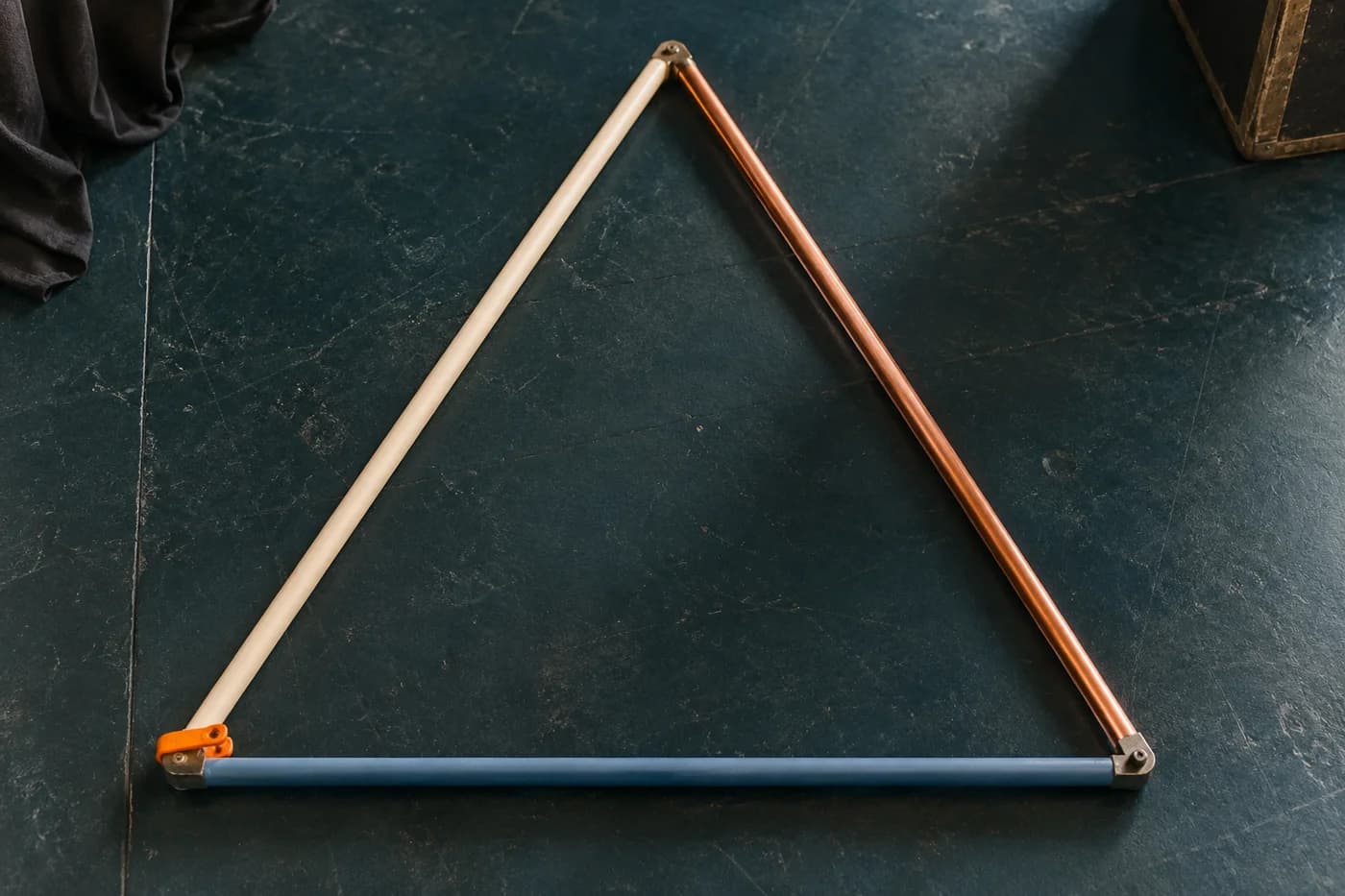 Vista cenital de un marco triangular formado por varillas marfil, cobriza y azul con una pinza naranja sobre un escenario verde azulado
